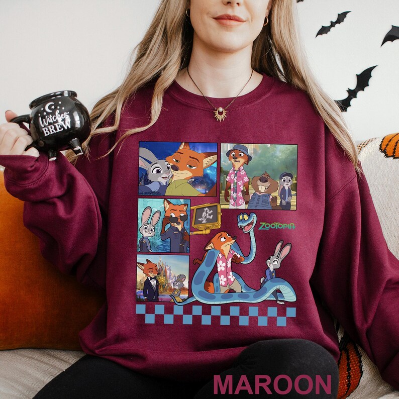 May include: Maroon sweatshirt featuring a colorful graphic of characters from the animated movie Zootopia. The design includes images of a fox, a rabbit, and other characters in a comic strip style. The word "Zootopia" is also visible.