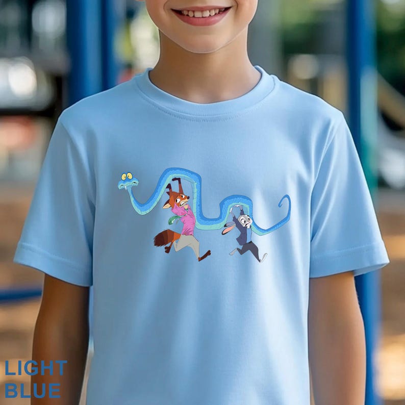 May include: A light blue short-sleeve t-shirt featuring a graphic print of a cartoon fox and rabbit running while being chased by a long, winding blue snake. The fox wears a pink shirt and the rabbit wears a dark blue jacket and pants.