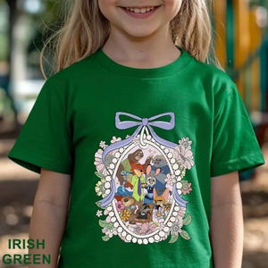 Pu&ograve; includere: T-shirt verde irlandese con un'illustrazione di personaggi di animali in un ovale, decorata con fiori e un fiocco lavanda. Le parole "Irish Green" sono stampate in basso sulla maglietta.