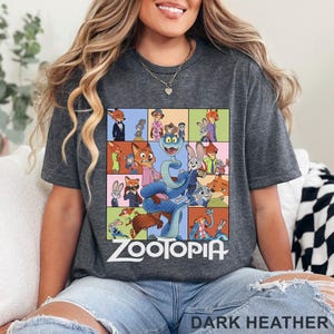 May include: A dark heather grey t-shirt featuring a grid of colourful Zootopia character illustrations, including Nick Wilde, Judy Hopps, and a large blue snake in the centre. The word Zootopia is printed in white text below the graphic design.