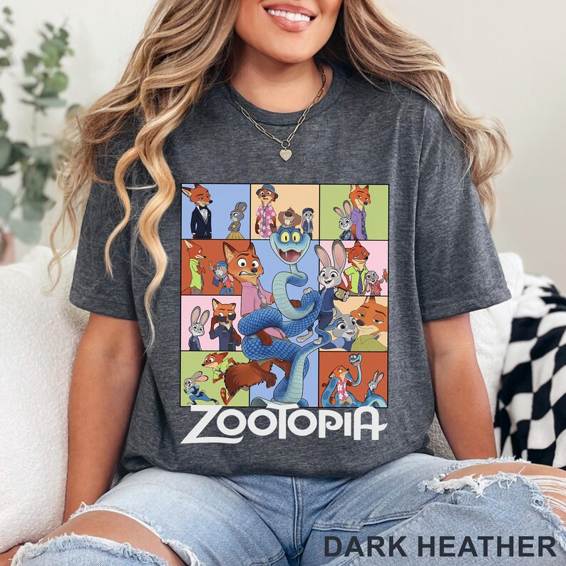 May include: A dark heather grey t-shirt featuring a grid of colourful Zootopia character illustrations, including Nick Wilde, Judy Hopps, and a large blue snake in the centre. The word Zootopia is printed in white text below the graphic design.