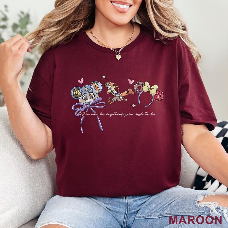 May include: A maroon short-sleeve t-shirt featuring a graphic print of a rabbit character with a police badge, a fox character, and two headbands. The text "You can be anything you wish to be" is printed in white cursive script below the illustrations.