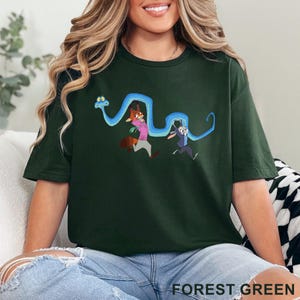 May include: A forest green short-sleeve t-shirt featuring a graphic print of a long blue snake winding around two cartoon characters, a fox in a pink shirt and a rabbit in a blue uniform, who are both running with expressions of surprise.
