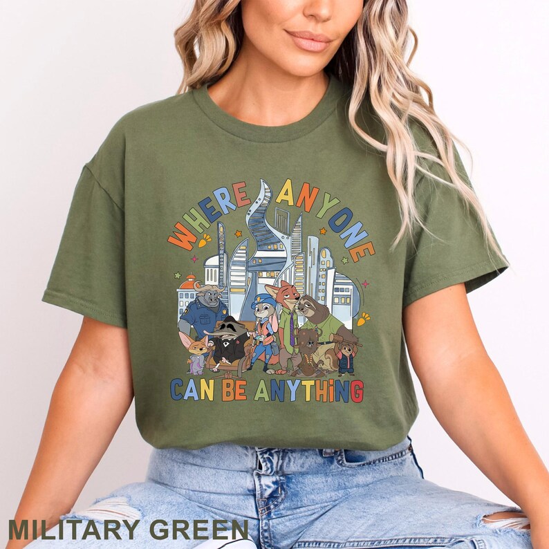 May include: Military green t-shirt featuring a colorful graphic with the text "WHERE ANYONE CAN BE ANYTHING" and cartoon characters. The design includes a cityscape and various animal characters. The shirt is a crew neck style.