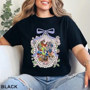Pu&ograve; includere: T-shirt nera con un'illustrazione a cartoni animati colorata di vari personaggi di animali all'interno di una cornice ovale, circondata da fiori e un fiocco. La parola "BLACK" &egrave; stampata in basso.