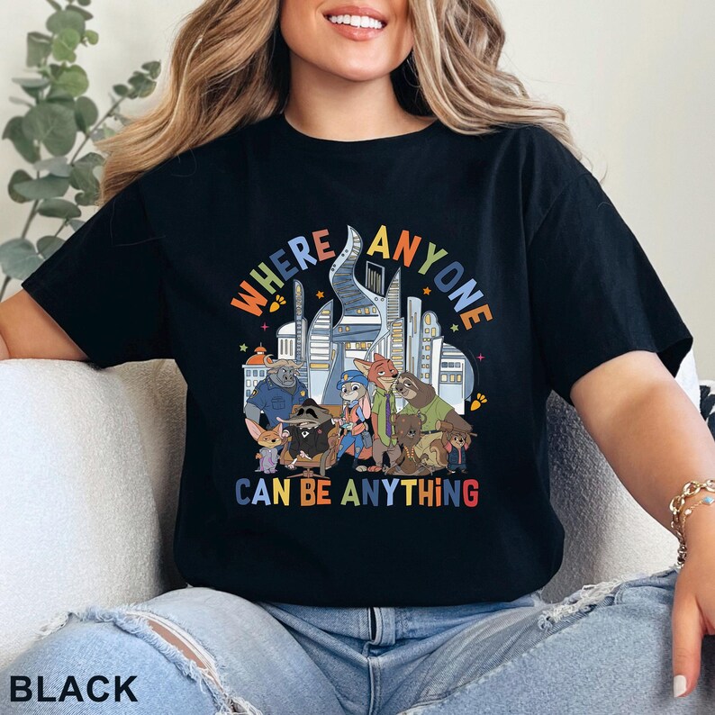 May include: Black t-shirt featuring a colorful graphic of cartoon animals and city buildings. The text on the shirt reads "WHERE ANYONE CAN BE ANYTHING". The design includes a variety of animal characters in a cityscape setting.