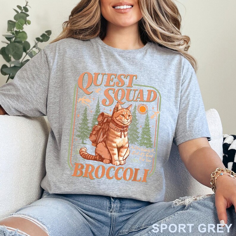May include: Sport grey t-shirt with a graphic featuring a cat wearing a backpack, the words "Quest Squad Broccoli", and a forest scene. The design is in orange, green, and light blue.
