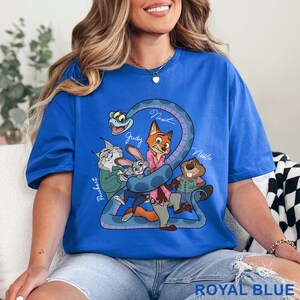 Puede incluir: Una camiseta de manga corta en color azul real con un estampado gr&aacute;fico de personajes animados, incluyendo un zorro, un conejo, un lince, un castor y una serpiente. El texto "Pawbert", "Judy", "Nick" y "Nibbles" aparece en letra blanca junto a cada personaje.