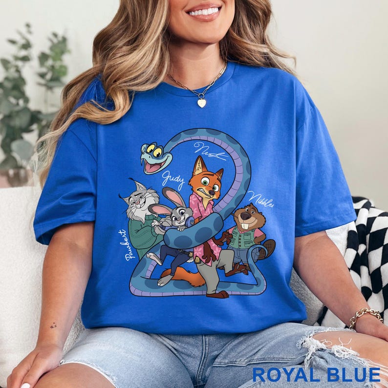 Puede incluir: Una camiseta de manga corta en color azul real con un estampado gr&aacute;fico de personajes animados, incluyendo un zorro, un conejo, un lince, un castor y una serpiente. El texto "Pawbert", "Judy", "Nick" y "Nibbles" aparece en letra blanca junto a cada personaje.