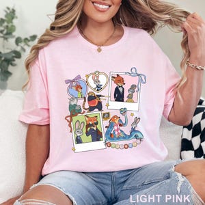 May include: A light pink t-shirt featuring a graphic print of Zootopia characters including Nick Wilde and Judy Hopps. The design includes multiple polaroid-style frames, heart-shaped borders, ribbons, and stars, with the text Zootopia at the bottom.