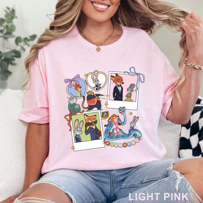 May include: A light pink t-shirt featuring a graphic print of Zootopia characters including Nick Wilde and Judy Hopps. The design includes multiple polaroid-style frames, heart-shaped borders, ribbons, and stars, with the text Zootopia at the bottom.