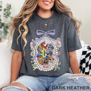 Pu&ograve; includere: T-shirt grigio scuro con un'illustrazione colorata di personaggi di animali dei cartoni animati all'interno di una cornice ovale ornata di fiori e un fiocco viola. Il testo "DARK HEATHER" &egrave; nella parte inferiore della maglietta.