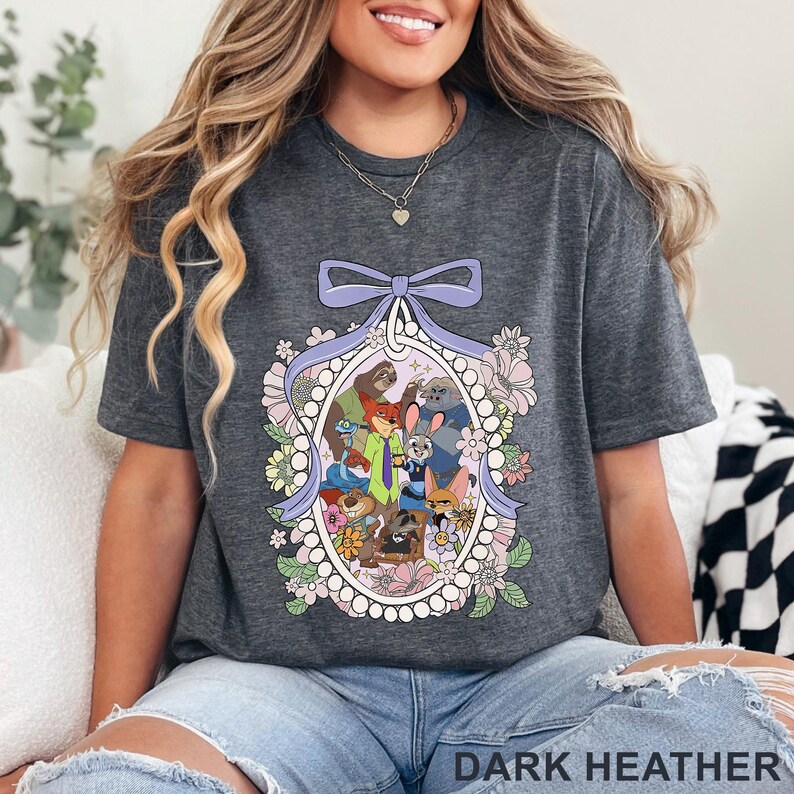Pu&ograve; includere: T-shirt grigio scuro con un'illustrazione colorata di personaggi di animali dei cartoni animati all'interno di una cornice ovale ornata di fiori e un fiocco viola. Il testo "DARK HEATHER" &egrave; nella parte inferiore della maglietta.