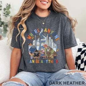 May include: Dark heather gray t-shirt featuring a colorful graphic with the text "WHERE ANYONE CAN BE ANYTHING" and cartoon characters in front of a city skyline. The shirt is a comfortable, casual style.