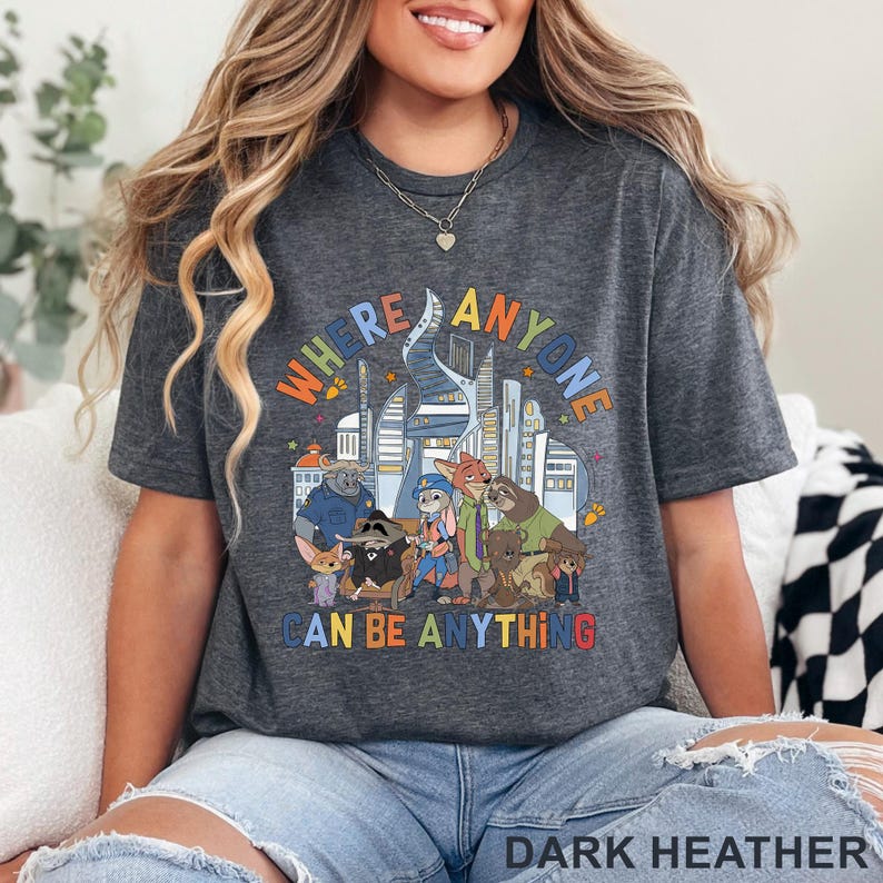 May include: Dark heather gray t-shirt featuring a colorful graphic with the text "WHERE ANYONE CAN BE ANYTHING" and cartoon characters in front of a city skyline. The shirt is a comfortable, casual style.