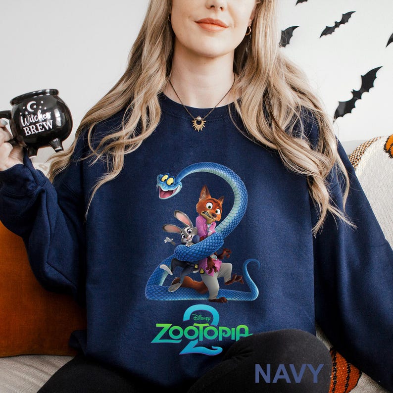 May include: Navy blue sweatshirt featuring a colorful graphic of characters from the Disney movie Zootopia. The design includes Judy Hopps, Nick Wilde, and a blue snake wrapped around the number 2. The word "Zootopia" is printed below the characters.