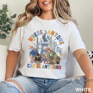 May include: White t-shirt featuring a colorful graphic with the text "WHERE ANYONE CAN BE ANYTHING" above a cartoon cityscape and animal characters. The shirt is made of a soft material.