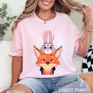 May include: Pale pink t-shirt with a cartoon fox and rabbit design. The fox is orange with a black jacket, and the rabbit is grey with long ears. The text "LIGHT PINK" is at the bottom of the shirt.