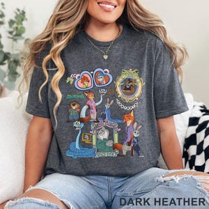 May include: Dark heather gray t-shirt featuring a colorful graphic of characters from the animated movie "Zootopia." The design includes various animal characters in detailed illustrations. The text "Zootopia" is also visible.