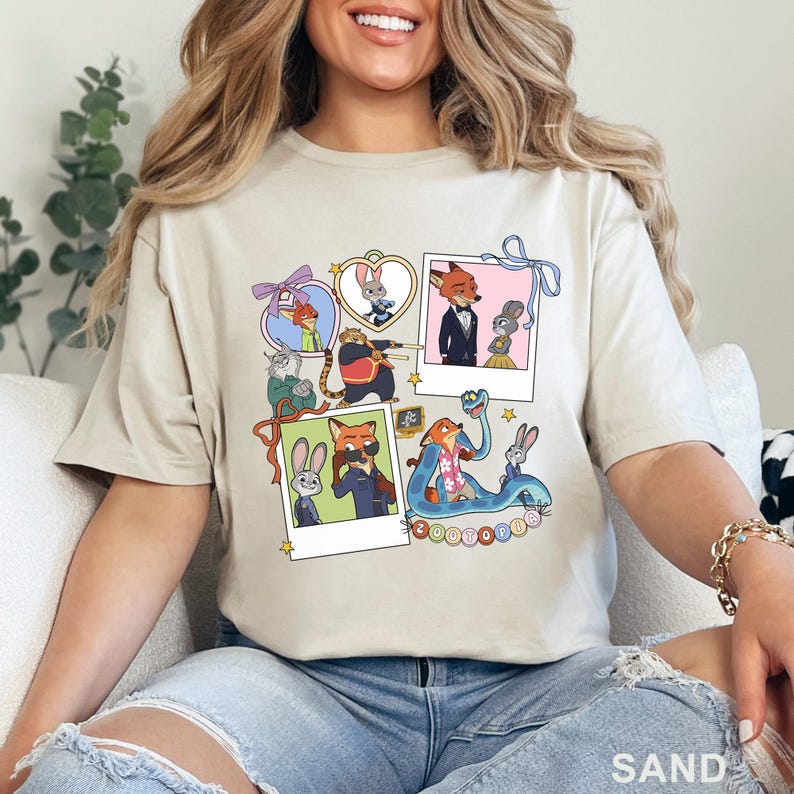 May include: A sand-colored t-shirt featuring a graphic print of Zootopia characters including Judy Hopps and Nick Wilde. The design includes multiple polaroid-style frames, heart-shaped borders, and a blue snake illustration with the text Zootopia.