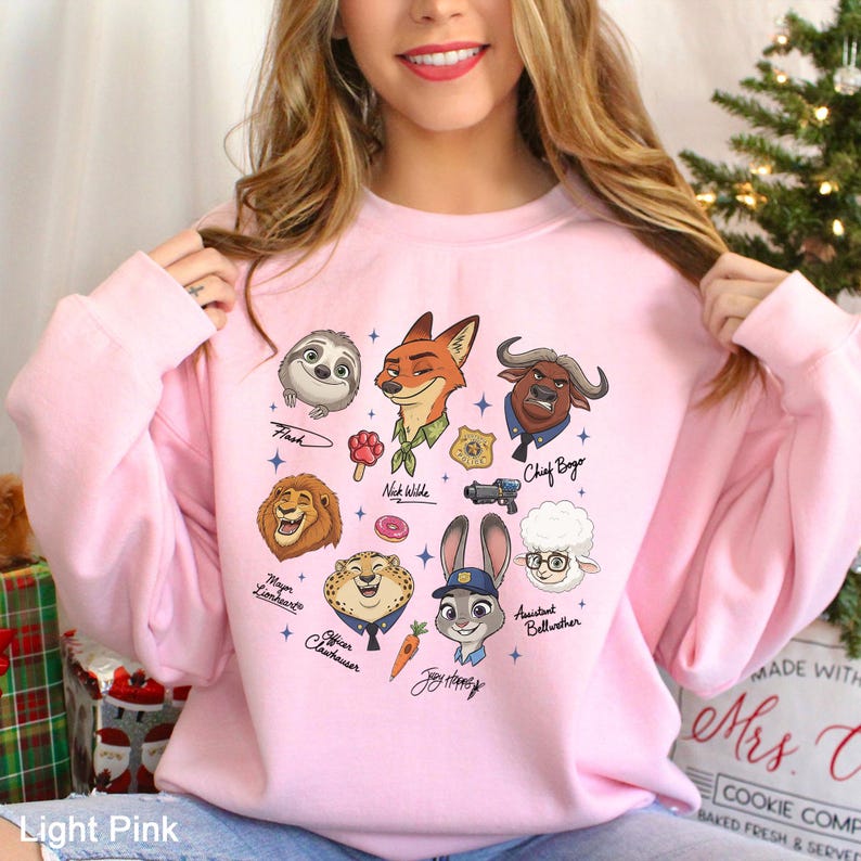 May include: Light pink sweatshirt featuring cartoon illustrations of characters from Zootopia, including Nick Wilde, Judy Hopps, and Chief Bogo. The design includes character names and various small graphics.