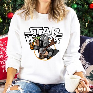 May include: White sweatshirt featuring a graphic of The Mandalorian and Grogu from Star Wars. The design includes the Star Wars logo and an orange circle around the characters. The Mandalorian is holding a weapon and Grogu is in a bag.