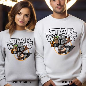 May include: Sport grey and white sweatshirts featuring a Star Wars design. The design includes the Star Wars logo, a cartoon Mandalorian character, and Grogu. The sweatshirts are long-sleeved.