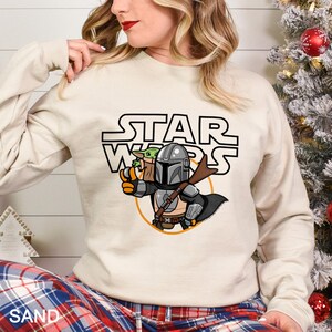 May include: Sand-colored sweatshirt featuring a Star Wars graphic. The graphic depicts the Mandalorian holding Baby Yoda, with the Star Wars logo above. The sweatshirt has long sleeves and a crew neck.