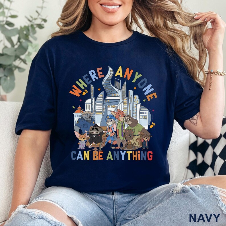 May include: Navy blue t-shirt with a colorful graphic featuring cartoon characters and the text "WHERE ANYONE CAN BE ANYTHING." The design includes a cityscape and various animal figures. The shirt is a comfortable, casual wear.
