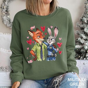 May include: Military green sweatshirt with a watercolour illustration of Nick Wilde and Judy Hopps from Zootopia, surrounded by pink hearts. Nick wears a green shirt and tie, Judy a police uniform. The text "MILITARY GREEN" is at the bottom.