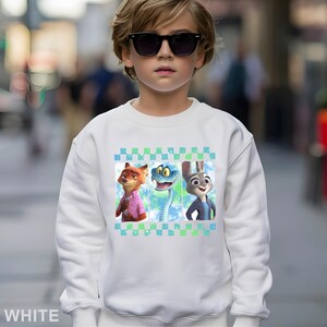 May include: White sweatshirt with a design of three cartoon characters: a fox in a pink shirt, a blue snake, and a rabbit in a blue jacket. The design is framed by a green and blue checkered pattern. The sweatshirt is worn by a child wearing sunglasses.