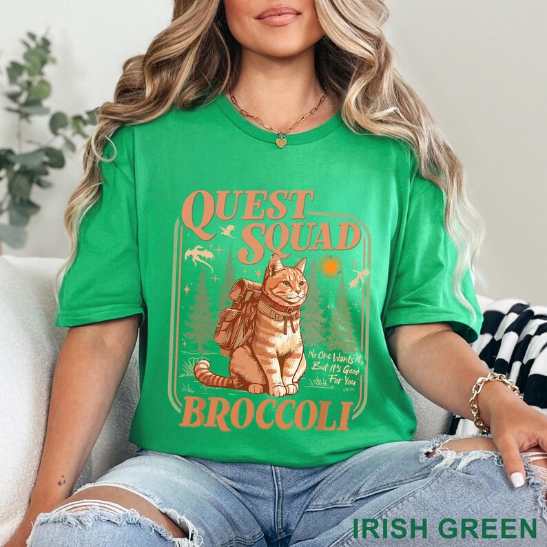May include: Irish green t-shirt with a graphic featuring a cat wearing a backpack, the words "QUEST SQUAD BROCCOLI", and the text "No One Wants It, But It's Good For You". The shirt is a crew neck.