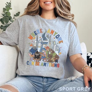 May include: Sport grey t-shirt featuring a colorful graphic of cartoon characters and a cityscape. The text on the shirt reads "WHERE ANYONE CAN BE ANYTHING". The shirt is made of a soft material.