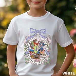 Pu&ograve; includere: T-shirt bianca con grafica a cartoni animati dei personaggi di "Zootropolis" all'interno di una cornice ovale, fiocco lilla e dettagli floreali. La parola "WHITE" &egrave; stampata in basso a destra. Una maglietta per bambini.