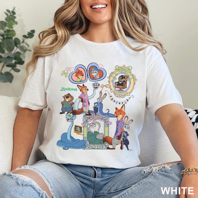 May include: White t-shirt featuring a colorful graphic of characters from the animated movie "Zootopia." The design includes Nick Wilde, Judy Hopps, and other characters in a cartoon style. The word "Zootopia" is also printed on the shirt.