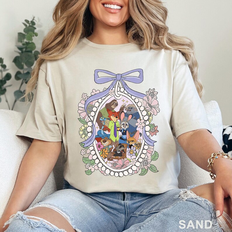 Pu&ograve; includere: T-shirt color sabbia con una grafica a cartoni animati colorata di vari personaggi di animali all'interno di una cornice ovale floreale con un fiocco viola. La parola "SAND" &egrave; stampata in basso a destra.
