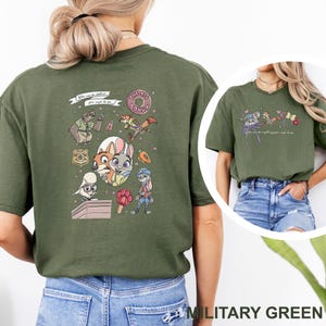 May include: An olive green t-shirt featuring a Zootopia-themed graphic on the back with characters like Judy Hopps and Nick Wilde, a donut, a badge, and a carrot. The front chest area displays a smaller graphic of character ears and text.