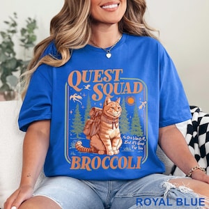 May include: Royal blue t-shirt with a graphic featuring a cat wearing a backpack, the words "QUEST SQUAD BROCCOLI", and the phrase "No One Wants It, But It's Good For You". The shirt is a solid color.