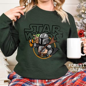 May include: Forest green sweatshirt featuring a cartoon illustration of The Mandalorian and Grogu from Star Wars. The design includes the Star Wars logo and a circular graphic. The sweatshirt is a solid color.