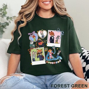 May include: A forest green short-sleeve t-shirt featuring a graphic print of various Zootopia characters, including Judy Hopps and Nick Wilde, arranged in a collage of polaroid-style frames, heart shapes, and ribbons with the text Zootopia.