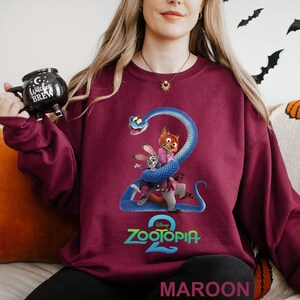 May include: Maroon sweatshirt featuring a graphic of characters from Zootopia 2, including a fox, rabbit, and snake. The Zootopia 2 logo is in teal. The word "MAROON" is printed at the bottom of the sweatshirt.