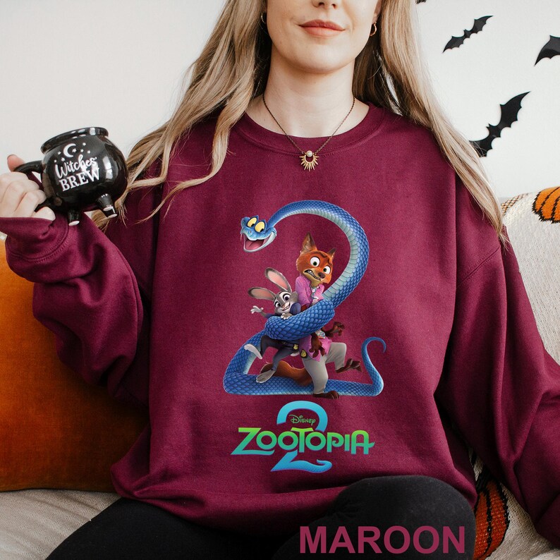May include: Maroon sweatshirt featuring a graphic of characters from Zootopia 2, including a fox, rabbit, and snake. The Zootopia 2 logo is in teal. The word "MAROON" is printed at the bottom of the sweatshirt.