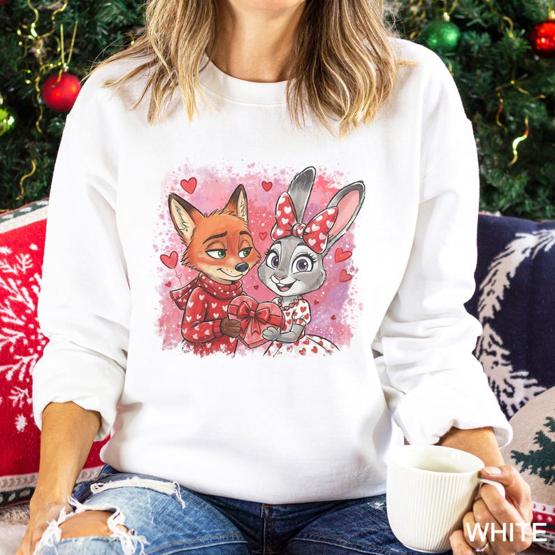 May include: White sweatshirt featuring a cartoon fox and bunny holding a heart-shaped gift. The fox wears a red scarf and the bunny has a bow with hearts. The design is set against a pink background with heart accents. A festive holiday theme.