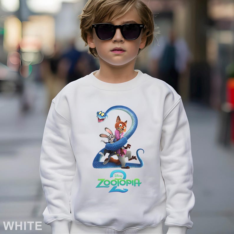 May include: White sweatshirt featuring a colorful graphic from the Disney movie "Zootopia 2." The design includes a blue snake, a fox, and a rabbit. The text "Zootopia 2" is printed in teal. The sweatshirt is worn by a person wearing sunglasses.