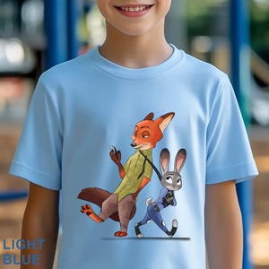 May include: Light blue t-shirt featuring a cartoon illustration of Nick Wilde and Judy Hopps from Zootopia. Nick, a fox, is holding a phone, while Judy, a rabbit, walks beside him. The text "LIGHT BLUE" is printed at the bottom.