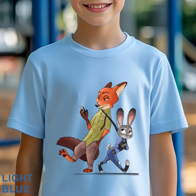 May include: Light blue t-shirt featuring a cartoon illustration of Nick Wilde and Judy Hopps from Zootopia. Nick, a fox, is holding a phone, while Judy, a rabbit, walks beside him. The text "LIGHT BLUE" is printed at the bottom.