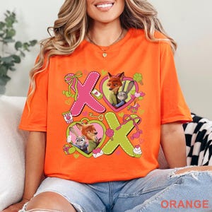 Peut inclure: Un t-shirt orange orn&eacute; d'un grand X rose et d'un O vert. Les lettres sont d&eacute;cor&eacute;es avec les personnages de Zootopie, Nick Wilde et Judy Hopps, &agrave; l'int&eacute;rieur de c&oelig;urs, entour&eacute;s de petites illustrations de bonbons, de n&oelig;uds et de c&oelig;urs.
