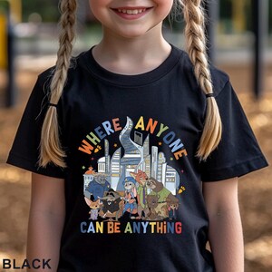 May include: Black t-shirt featuring a colorful graphic with the text "WHERE ANYONE CAN BE ANYTHING." The graphic includes a cityscape and cartoon characters. The shirt is made of a soft material.