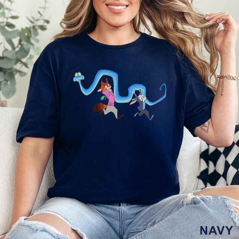 May include: A navy blue short-sleeve t-shirt featuring a graphic print of a long, winding blue snake held up by a cartoon fox wearing a pink shirt and a cartoon rabbit wearing a blue uniform. The design is centered on the chest of the garment.