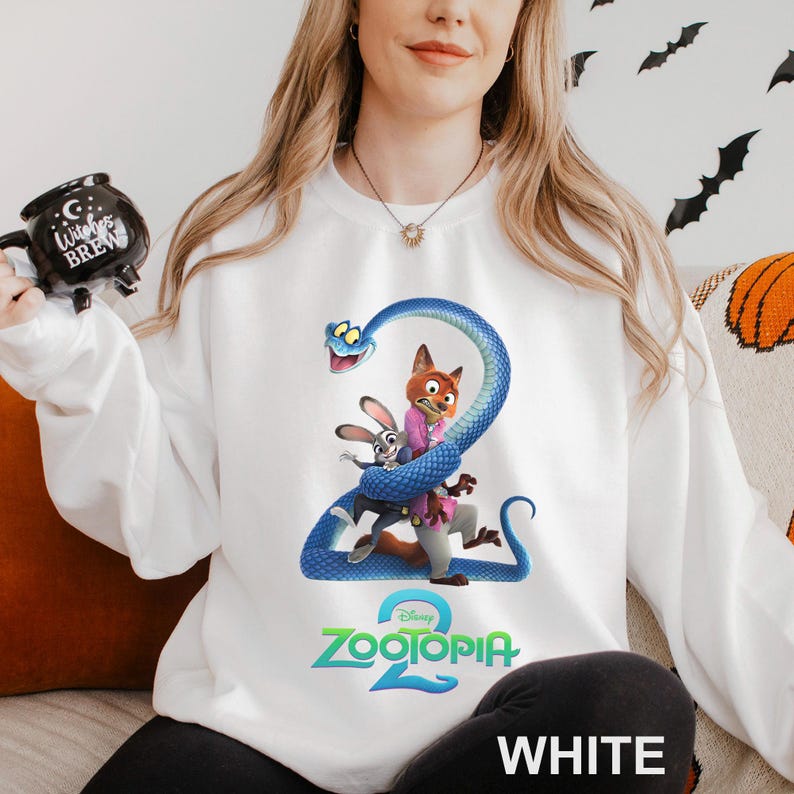 May include: White sweatshirt featuring a colorful graphic of characters from the movie Zootopia 2. The design includes a blue snake wrapped around a fox and a rabbit. The text "Zootopia 2" is displayed in green and blue.